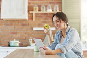 Healthy food and lifestyle concept. Pretty female eats apple, looks for new diet in internet, uses tablet computer, being glad to loose weight. Woman watches film on kitchen as dinner is prepared