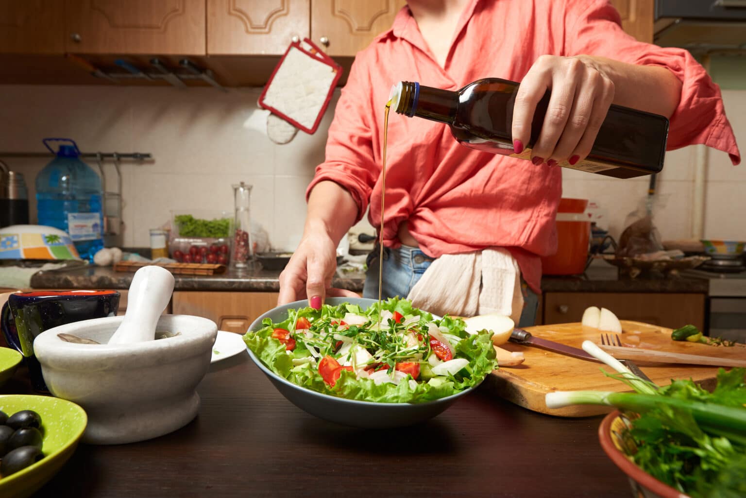 Woman adding olive oil into healthy greek salad. Kitchen on background