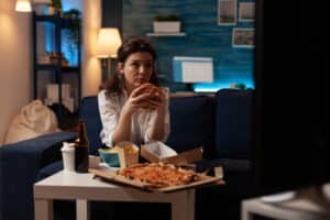 Woman sitting on sofa eating tasty delicious burger watching news on television in front of coffee table with large pizza. Caucasian female enjoying takeaway junk-food order.
