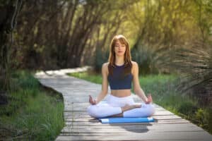 Young woman doing yoga in nature. Female wearing sport clothes in lotus figure. Girl sitting on wooden road.