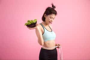 a picture of a woman on a pink background measuring her waist size and holding a bowl of salad on her lazy keto journey