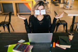 young pretty tired woman with paper stickers on glasses sitting at table in black shirt working on laptop in co-working office, funny face emotion, problem, workplace, holding hands up